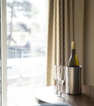 Bright hotel room with a round table, wine bottle in ice bucket, two glasses, and large window view. Relaxed luxury stay.