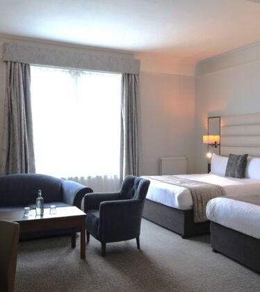 Spacious hotel room with two double beds, cosy seating area, flat-screen TV, natural light, modern decor.