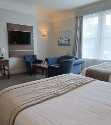 Elegant hotel room with two beds, cosy seating area, desk, large window with curtains, wall-mounted TV, and coffee amenities.