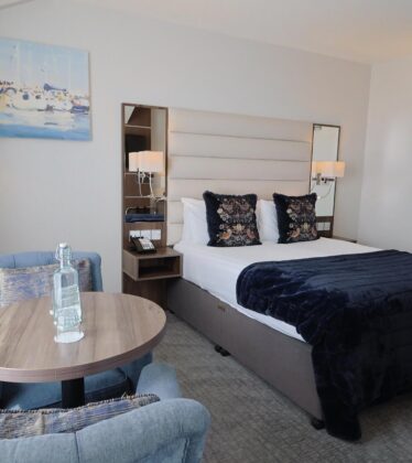 Elegant hotel room with king-sized bed, plush pillows, soft armchairs, and a scenic marina artwork, ideal for relaxation.