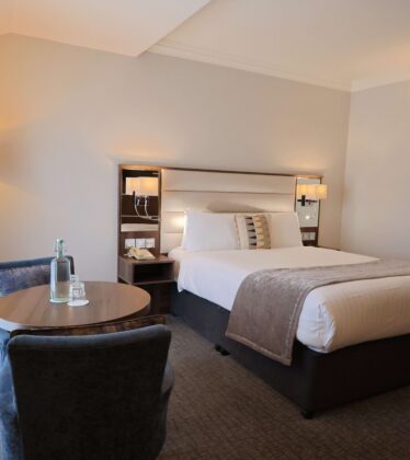 Spacious hotel room with king-size bed, cosy armchairs, natural light, luxury decor, and modern lighting.