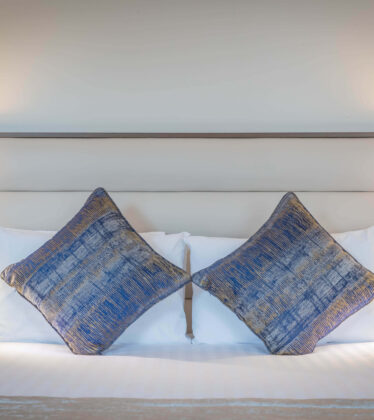 Spacious hotel room with plush bed, elegant blue cushions, warm lighting, and large windows offering natural light.