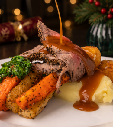 Roast beef with pepper crust, gravy pouring over mashed potato, roast carrots, crispy potatoes and broccoli in festive hotel dining.