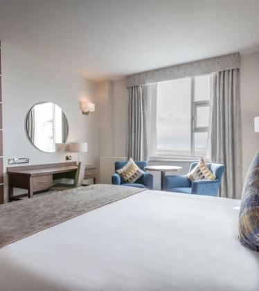 Bright double hotel room with king bed, TV, desk, seating area by window and sea view, modern lamps and soft linens